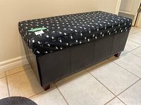 View of the front and side of the rectangular pleather storage ottoman covered by a fitted black cover with white and gray spot pattern, showing wooden block feet and tile floor.
