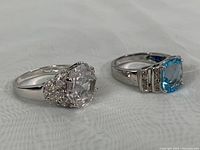 Side angle photo of both 925 silver rings showing faceted stones and prong settings on a white fabric background.