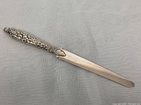Full view of the sterling silver letter opener showing the polished blade and decorative floral handle on a textured light fabric background.