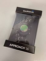 Front view of Garmin Approach S6 watch in original sealed box showing watch face with golf course interface displayed.