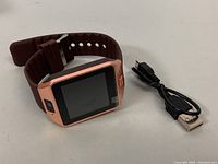 Rose gold square smart watch with brown wristband, next to black USB charger cable