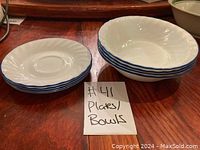 Photo showing 4 stacked white plates with blue rims and 4 stacked white bowls with blue rims on a wooden surface.