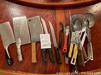 Wide shot showing assortment of kitchen utensils including cleavers, knives, peelers, ladles, and other miscellaneous tools on a wooden surface