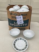 Cardboard box containing multiple white bowls and two white plates with a black geometric pattern around the edge, displayed outside on a table.