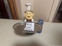 Front view of a ceramic or resin chef figurine holding a round metal serving tray, next to a water bottle for scale.