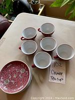 Photo of six Chinese tea cups with red and floral exterior and white inside, arranged near five matching plates.