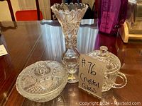Three pieces of crystal glassware displayed on a wooden table: a tall vase and two covered bowls.