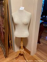 Front view of a 44 inch tall beige fabric covered female dress form mannequin on wooden tripod stand, photographed against a beige wall in a home interior.