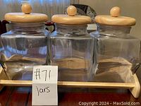 Front view of three clear glass jars with wooden lids on wooden frame stand, label '#71 jars' in foreground