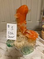 Front view of orange glass koi fish statue with flowers and leafy base on table.