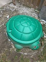 Green turtle-shaped sandbox closed with a lid, placed outdoors on grass and dirt.