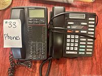 Pair of two black landline phones placed side by side, one Nokia model with numeric keypad and LCD screen and the other a multi-line office phone with numerous buttons, both with coiled cords and connected handsets.