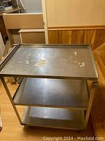 Metal restaurant cart featuring three shelves with raised edges and caster wheels, shown from side angle.