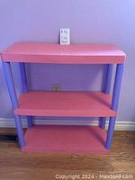 Photo showing a three-tier shelving unit with pink shelves and purple legs against a purple wall, placed on a wooden floor.