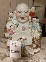 Porcelain Buddha statue with five children, showing color and detail on the figurines and garments. Statue placed on marble surface with sign labeled '#2 Buddha'.