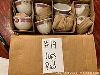 Eight porcelain tea cups packed in a box with dividers and paper padding to keep them separated. Each cup has red geometric patterned rim and floral details.
