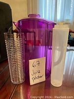 Photo showing a purple translucent plastic juice pitcher with lid, a tall clear plastic container with white lid, and a clear cylindrical container with spiral design, all marked with a note '67 Juice pitcher'.