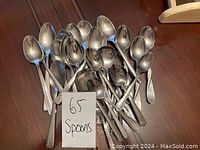 Photo of 25 assorted metal spoons in a pile on a wooden surface with a handwritten sign showing '65 Spoons'.