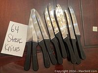 Photo of 10 serrated steak knives with black plastic handles arranged on wooden surface next to a handwritten sign reading '64 Steak Knives'.