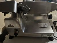 Side view of Omas meat slicer showing stainless steel body and brand label on bottom corner.