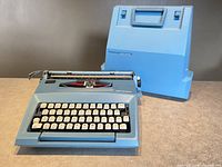 Front and side view of sky blue Smith Corona Courier typewriter with attached carrying case lid behind it, showing keyboard layout and general condition.