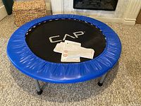 Front view of the assembled round fitness trampoline with blue padded edges and CAP logo on jumping mat, assembly instructions placed on mat.