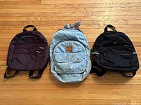 Front view of three backpacks: maroon-purple Roots sling backpack on the left, vintage denim Cricket backpack in the center, and black Roots sling backpack on the right.