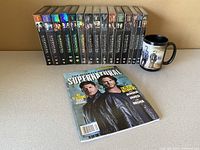 Photo showing 15 DVD box sets for seasons 1-15 of Supernatural, a Supernatural guide magazine, and a themed mug.