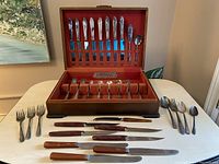 Full view of the flatware set arranged on the table outside of the wooden case, showing knives with Bakelite handles, silver forks, spoons, and some serving utensils.