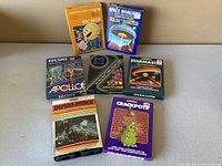 Seven vintage Atari video game cartridges with original boxes arranged on a table: Pac-Man, Space Invaders, Infiltrate Apollo, Lock'N'Chase, Starmaster, Crackpots, and Demon Attack.