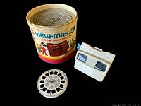 Photo showing the original cylindrical colorful box with Disney-themed graphics, a white View-Master viewer, and one reel on black background.