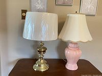 Photo showing both brass and pink painted ceramic vintage lamps side by side on a wooden table against a wall with framed pictures.