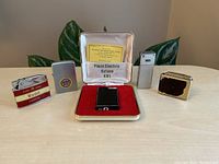 Wide view of five vintage lighters displayed with plants in background, showing variety of styles and brands