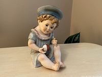 Front view of the bisque porcelain baby figure seated, wearing a large sailor hat and holding a ball, showing detailed hand-painted features and clothing with soft pastel colors.