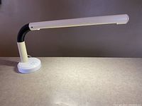 Side angled view showing the lamp fully extended with the white tubular light head and the black adjustable section near the circular white base with controls.