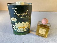 Photo showing White Nymph Luxury Talc Powder in a decorative metal tin with floral design and the White Shoulders cologne bottle with a pink round cap, both placed on a flat surface.