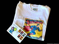 Close-up photo of folded white Roots Superman T-shirt with stamp sheet placed partially on top