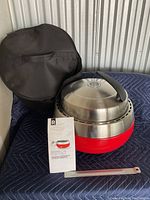Portable barbecue grill with lid, red base, black carrying bag, instruction manual, and metal tool on a quilted surface.