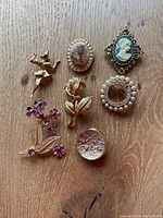 Photo of eight vintage brooches and pins on wooden surface showing variety of designs including cameo, flowers, and dancer.