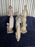 Photo showing three Lladro porcelain figurines including a standing woman, a man with fish, and a woman playing a flute on a quilted blue backdrop.