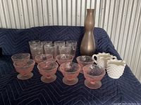 Assorted glassware including pink dessert dishes, clear tumblers, white hobnail pitchers, and a tall bronze vase arranged on a dark fabric background.