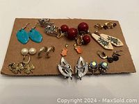 Various pairs of clip-on and pierced earrings laid out on cardboard showing different colors and styles including turquoise, red beads, leaves, pearls, and floral designs.
