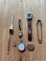 Top down photo of assorted watches, earrings, bracelet segment, compact mirror, and large safety pin laid out on a wooden surface