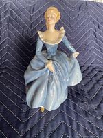 Porcelain figurine of a seated woman in blue dress shown from the front on a dark quilted fabric.
