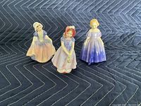 Three Royal Doulton porcelain figurines Ivy, Dinky Do, Marie shown together on dark padded cloth background.