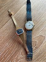 Two wristwatches on wooden surface: one rectangular gold-tone Timex; one round gold-tone Gruen with blue mesh strap