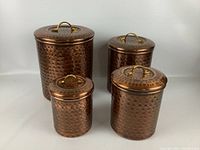 View of four copper hammered canisters with lids, displayed from front, showing nesting sizes and brass handles.