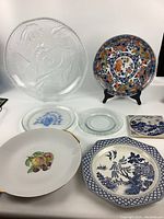 Lot of assorted decorative plates on white background including clear embossed glass platter and colorful floral plate