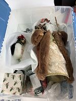 Plastic bin filled with assorted Christmas ornaments and two Santa figurines, some wrapped in tissue paper for protection.