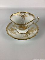 White and gold floral patterned tea cup with scalloped edge and gold trim. Ornate handle with gold accents.